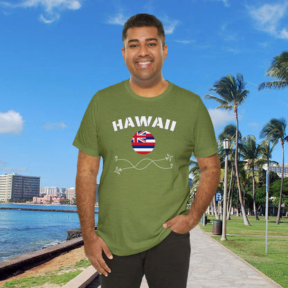 Brown skin man wearing a green cotton T-shirt featuring the word 'Hawaii' and its rounded flag, in a waterfront park background