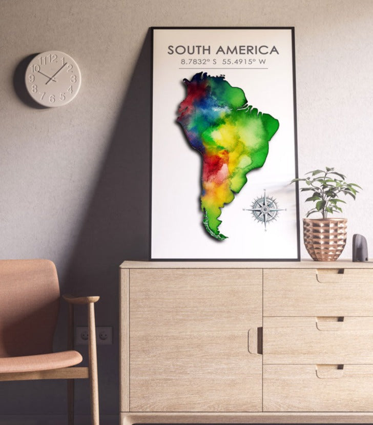 Black-framed colorful South America map poster on a light beige cabinet against a light pink wall with surrounding decor